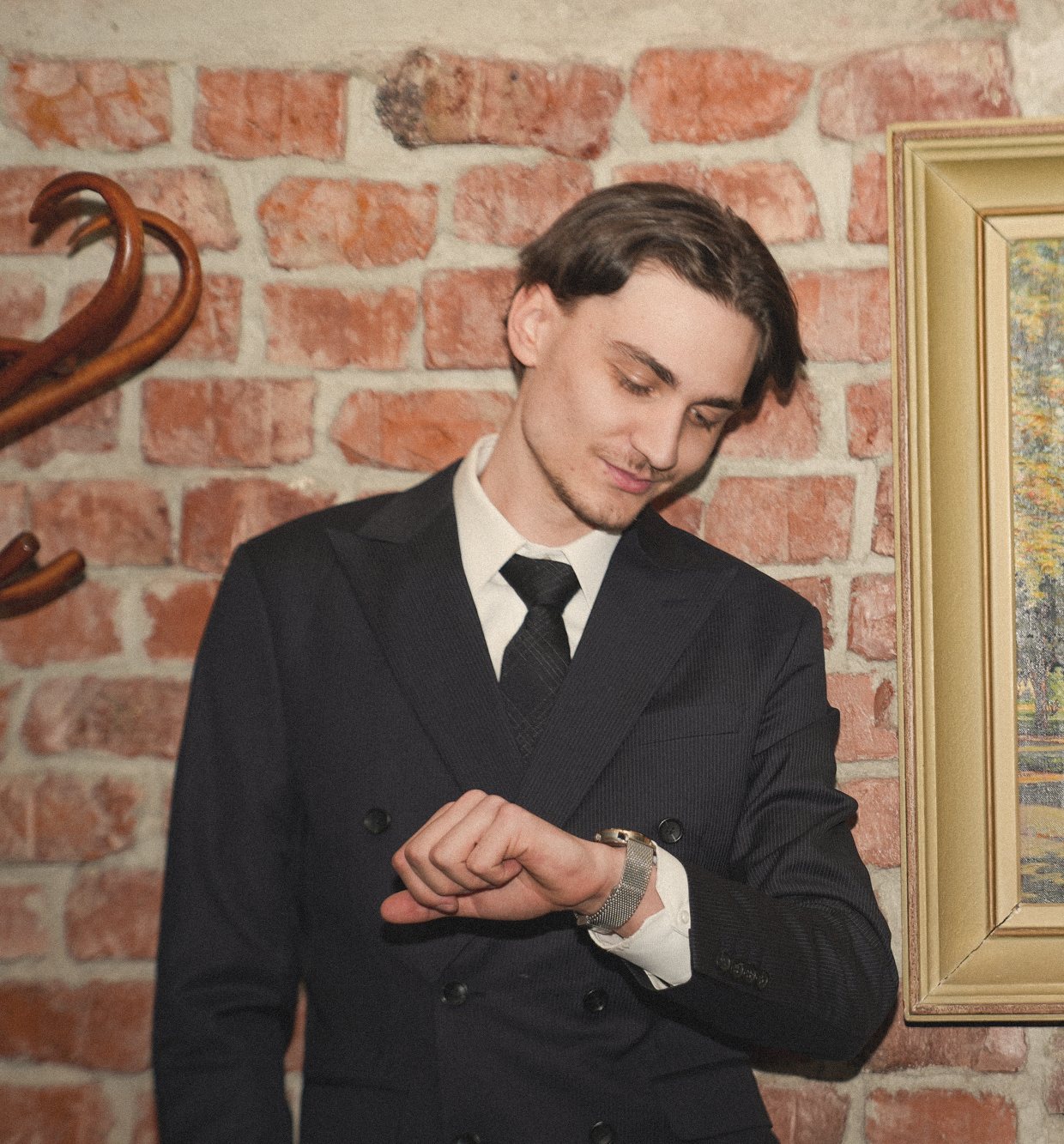 A man in a dark suit checking his watch in front of a brick wall